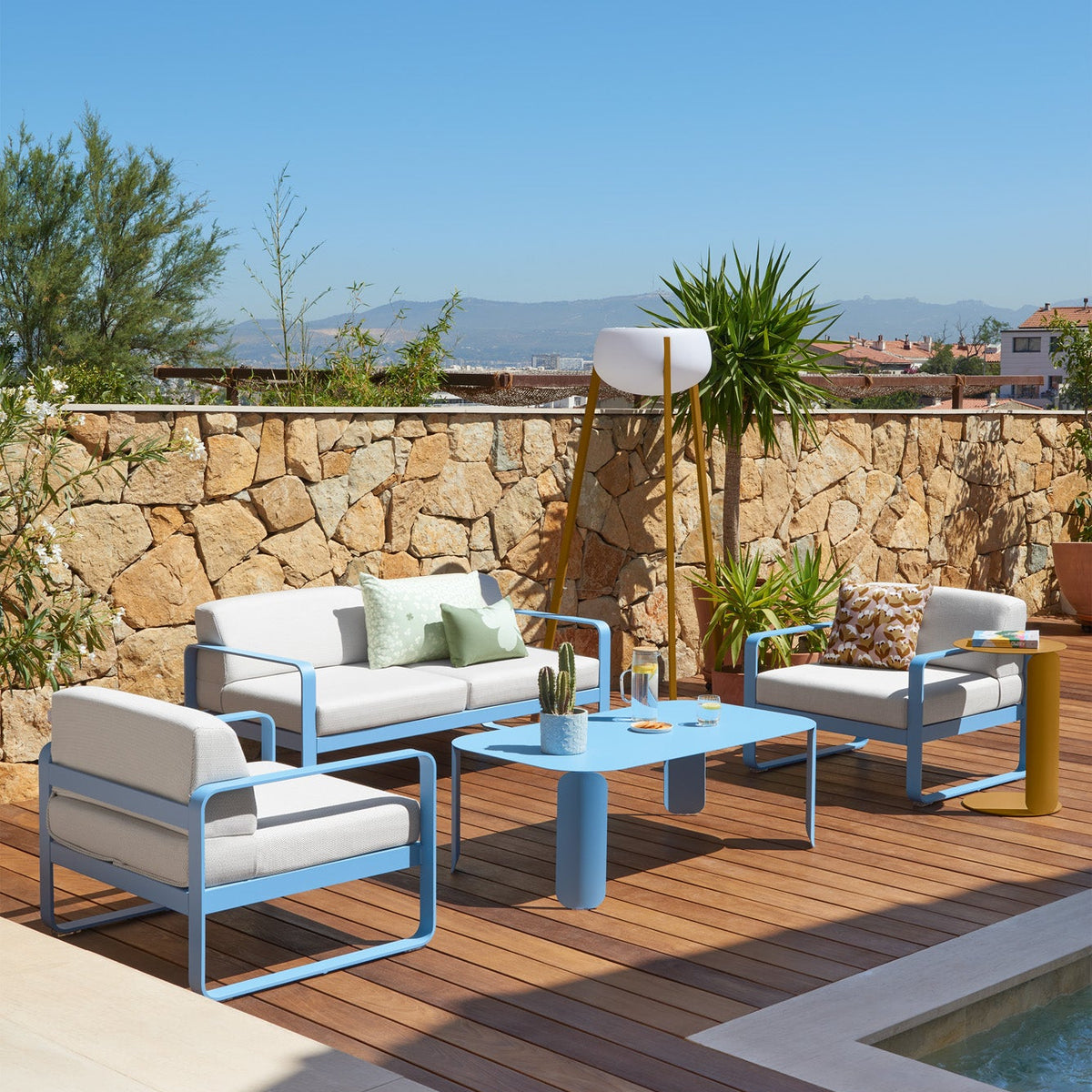 Outdoor patio set with blue chairs and table on a wooden deck with a stone wall and pool in the background.