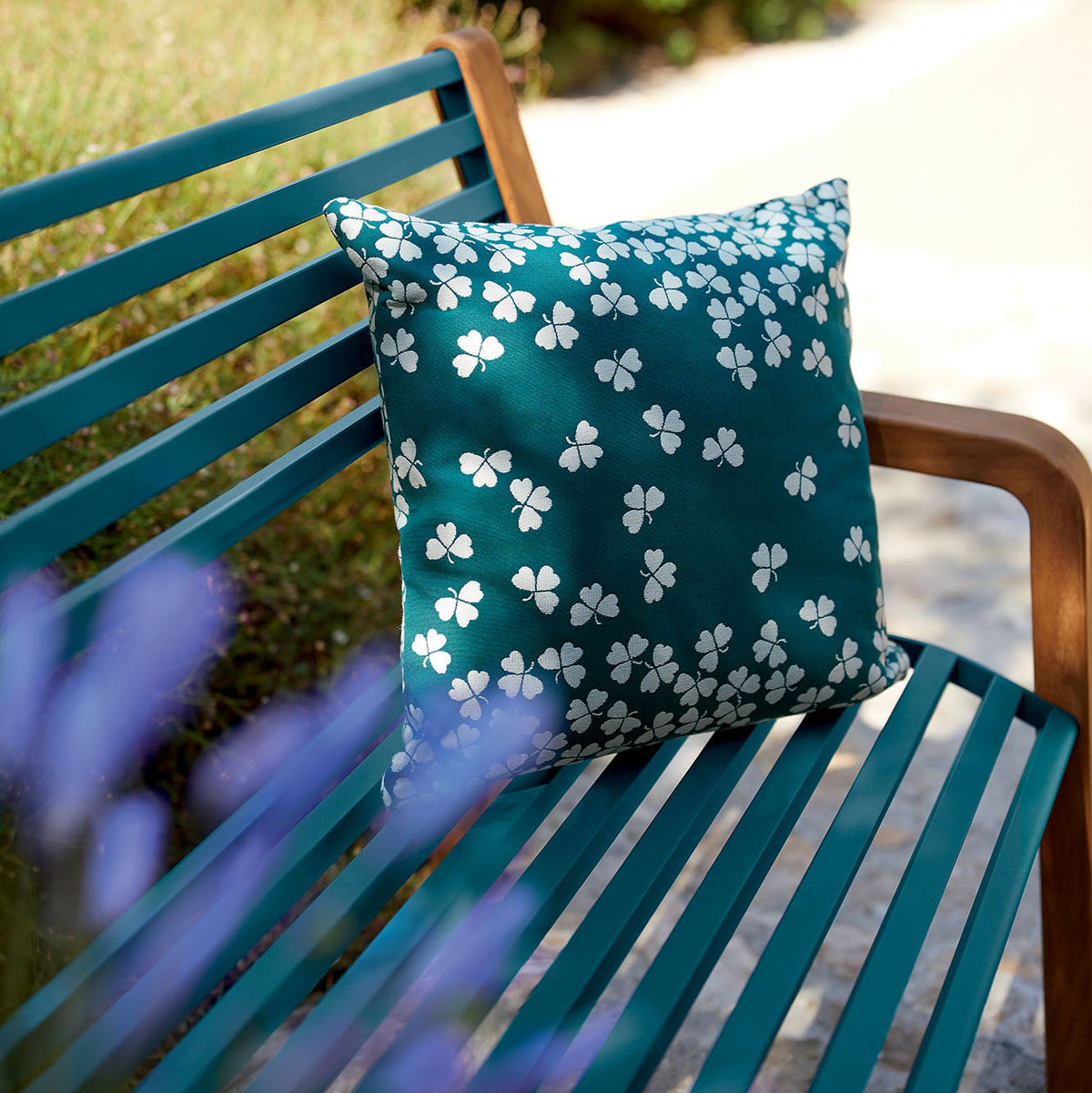 Teal bench with a decorative pillow featuring white floral patterns, set against a garden background.