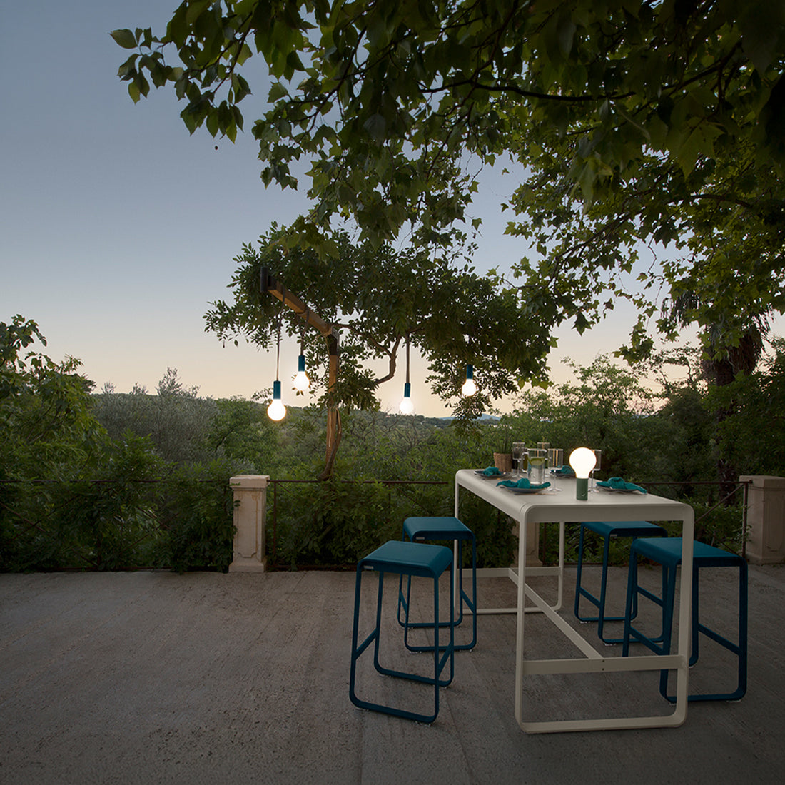 Outdoor bar table and stools under a tree at dusk.