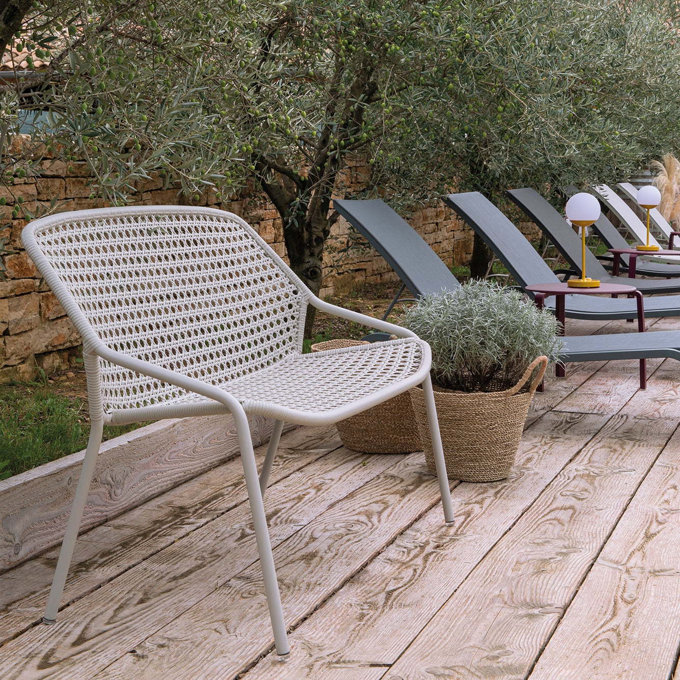 White wicker chair on a wooden deck with olive trees and lounge chairs in the background.