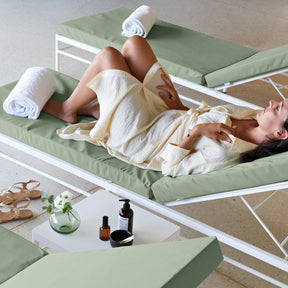 A woman relaxes on a white-framed outdoor lounge chair with a sage green cushion, wrapped in a cream towel. Another matching lounge chair sits behind her with rolled white towels, and a small side table holds skincare bottles and a glass vase with flowers.