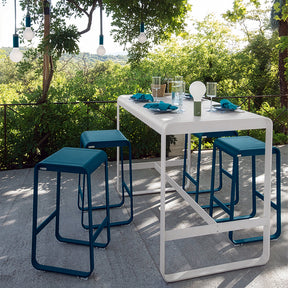 Outdoor dining setup with a bar table and blue stools on a patio with greenery in the background.